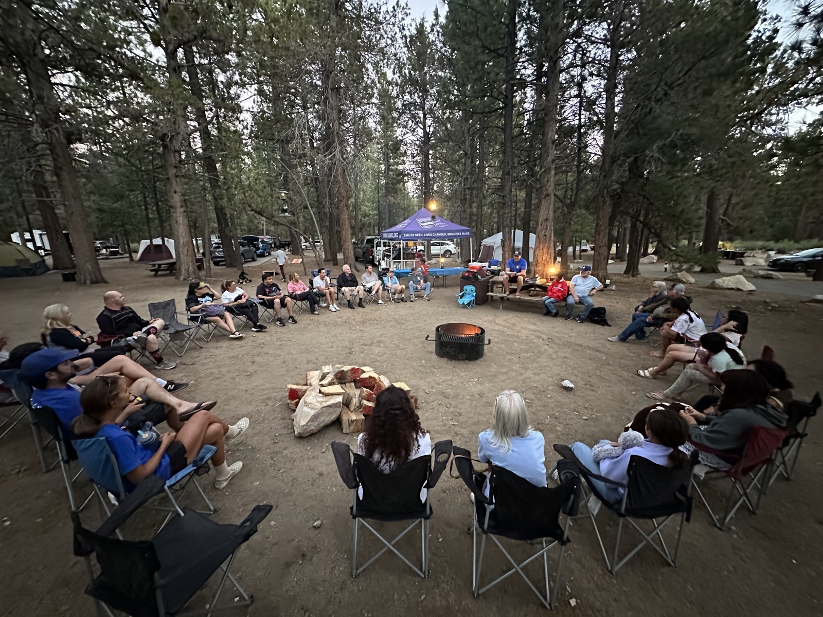 Campers sit in a circle of camp chairs around the fire ring under the pines at the overnight session.