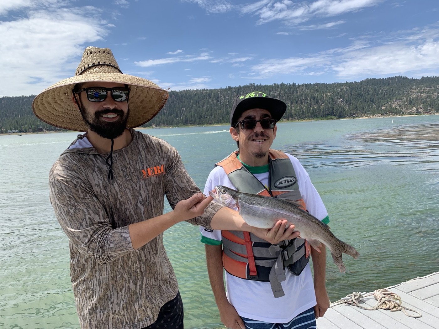A man in a straw hat holds up a freshly caught fish beside a young participant in a life jacket on the dock.