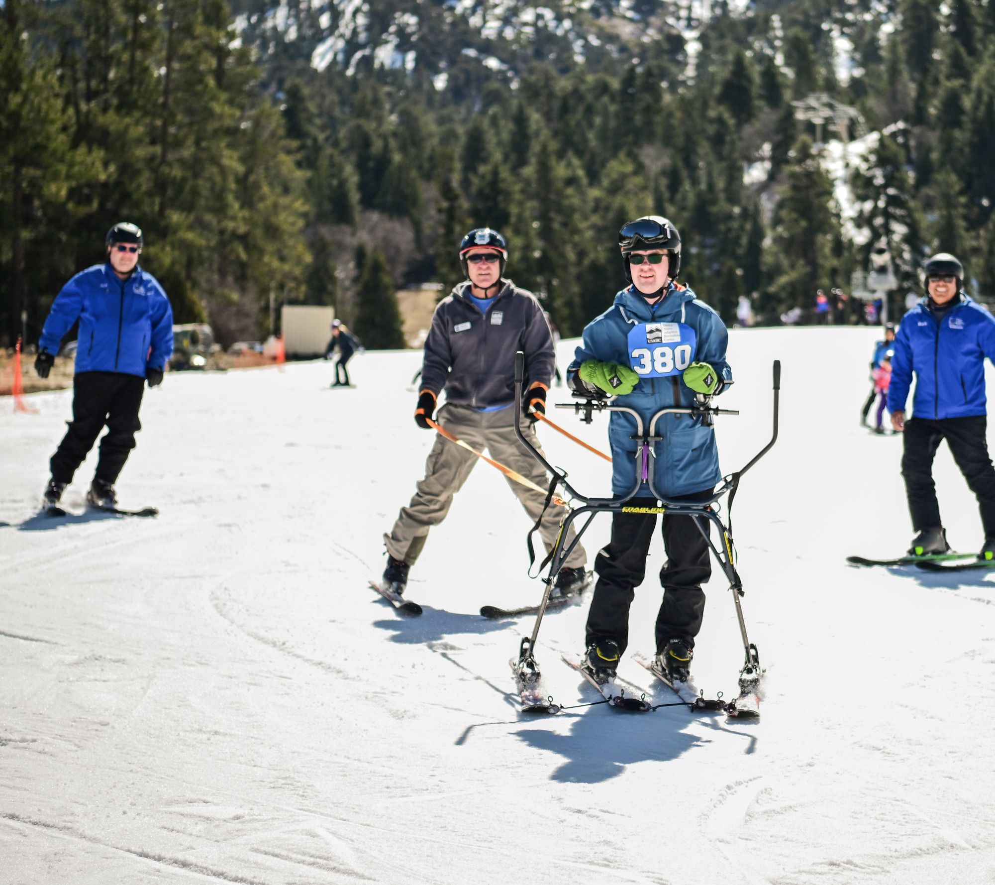 A participant in a slider frame skis toward the camera, surrounded by three USARC adaptive instructors.