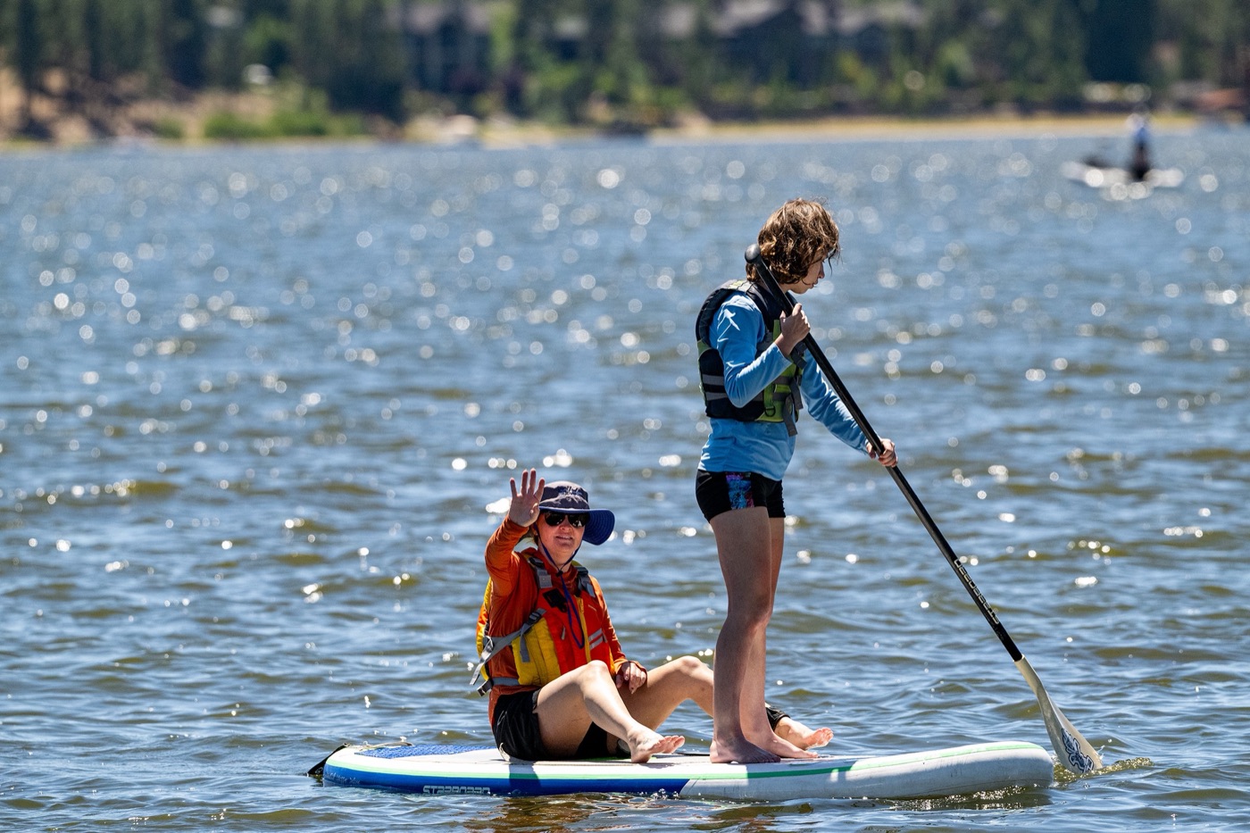 A young participant in a life jacket waves from a seat on a paddleboard while a volunteer stands and paddles.